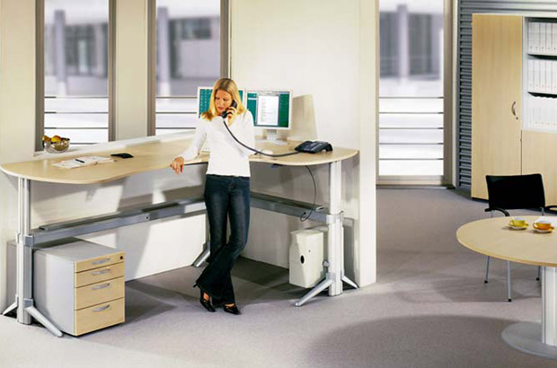 Frau steht telefonierend im Büro am verstellbaren Steh-Schreibtisch